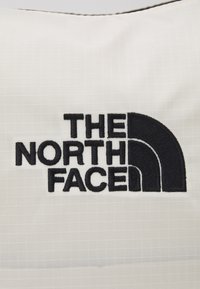 Biały materiał z siatkową teksturą ma wyraźne czarne haftowane logo z napisem "THE NORTH FACE" obok czarnego półokrągłego wzoru.