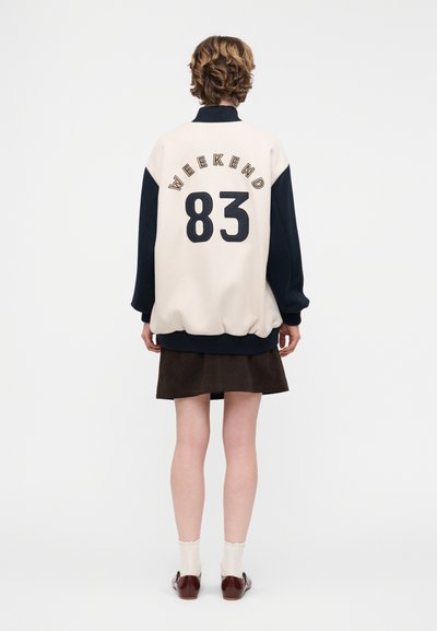 Άτομο με κοντά σγουρά μαλλιά, φοράει ένα διχρωμο φαρδύ πουλόβερ με την επιγραφή «WEEKEND 83» στην πλάτη, μια σκούρα φούστα, λευκές κάλτσες και καφέ παπούτσια.