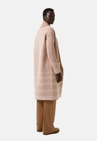 Woman wearing a pink and white textured long coat, brown wide-leg pants, and beige shoes, standing and looking over her shoulder.