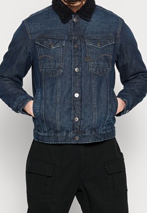 Giacca di jeans di colore blu scuro con colletto in shearling nero testurizzato, dotata di tasche frontali con bottoni e design aderente.