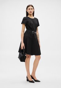 Moteris juoda dryžuota suknele su diržu, laiko juodą rankinę, vilki juodus aukštakulnius, stovi priešais paprastą foną.