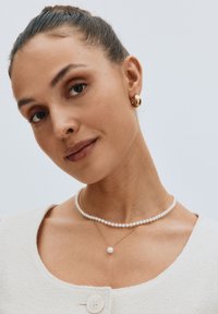 Goldene Ohrstecker, geschichtete Perlenketten in Weiß und Gold, strukturierte Cremebluse mit Rundhalsausschnitt und Knopfverschluss.