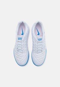 Baltos sintetinės futbolo bateliai su siuvinėtu tekstūru, su mėlynomis akcentais ir Nike logotipu. Su raišteliais ir paminkštinta apykakle patogumui.