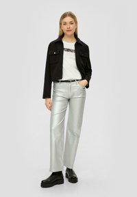 Zwarte cropped jas, witte T-shirt met grafische print, glanzende zilveren broek met brede pijpen, zwarte chunky laarzen, model staat met haar handen in de zakken.