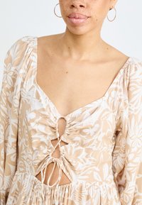 Robe à motifs floraux en beige et blanc, avec un décolleté en V, des manches bouffantes, des détails avec cordon à la taille, et une coupe ample.