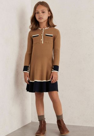 Brązowa sukienka z prążkowanego materiału, z rozszerzanym granatowym dołem, guzikami i kontrastującymi detalami kieszeni. W zestawie z brązowymi botkami i szarymi skarpetkami z dzianiny.