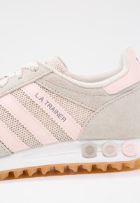 Světlé šedé tenisky s růžovými proužky a síťovaným svrškem, mají zaoblenou špičku, texturované semišové doplňky a bílou podrážku s barevnými knoflíky.