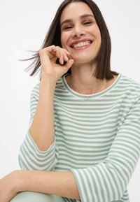 Grünes und weiß gestreiftes Langarmshirt mit rundem Halsausschnitt aus weichem Stoff. Auffällige silberne Halskette und lächelnder Gesichtsausdruck.