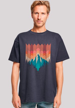 Navyblaue Baumwoll-T-Shirt mit einem geometrischen Berggrafik in Rot, Orange und Türkis, vor einem Sonnenuntergangshintergrund.