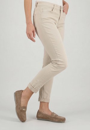 Kobieta ubrana w beżowe podwinięte spodnie i brązowe zamszowe mokasyny, stojąca z jedną stopą lekko uniesioną na jednolitym tle.