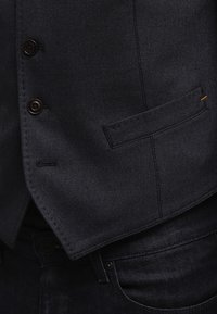 Mörkblå väst i texturerad tyg, med tre bruna knappar och en sidficka med orange accent. Bärs över mörka jeans.