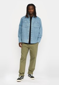 Lichtblauwe denimshirt met twee voorzakken, gedragen over een zwart topje, gecombineerd met olijfgroene broek en zwart-witte sneakers.