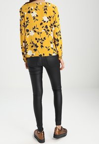 Blouse jaune à manches longues avec un motif floral blanc et noir, associée à un pantalon en simili cuir noir et des chaussures à enfiler marron.