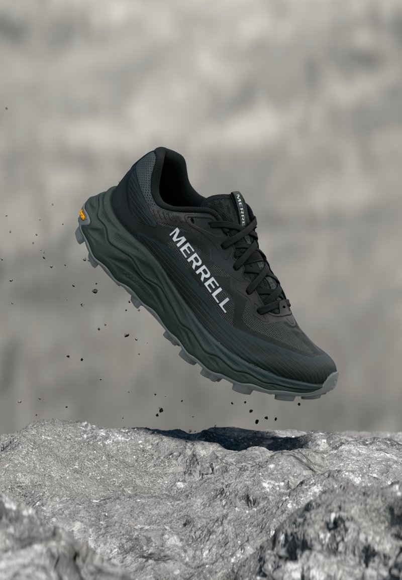 Μαύρο παπούτσι για τρέξιμο σε μονοπάτι Merrell με παχιά σόλα που αιωρείται πάνω από πετρώδες έδαφος, σωματίδια χώματος αιωρούμενα στον αέρα.
