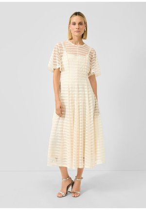 Vrouw draagt een crème midi-jurk met horizontale doorschijnende strepen en fladdermouwen, gecombineerd met metalen sandalen met hoge hakken, staand tegen een effen achtergrond.