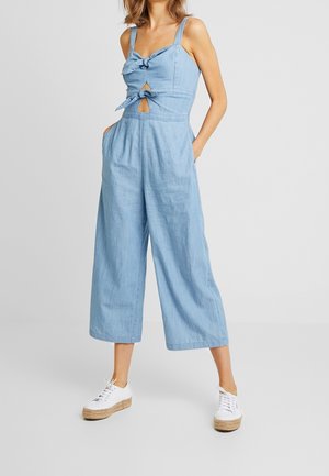 Kvinde iført lyseblå ærmeløs jumpsuit med bindedetaljer foran og vidde, cropped bukser, kombineret med hvide espadrillesneakers.