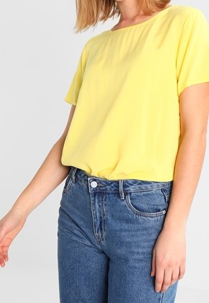 Osoba ubrana w jednolitą, żółtą koszulkę z krótkim rękawem, wciśniętą w wysokowcieniowe niebieskie dżinsy, stojąca na jasnym tle.