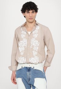 Camicia button-up beige chiaro con ricami floreali bianchi sul davanti. Presenta un colletto ampio e un tessuto testurizzato. Abbinata a jeans blu.