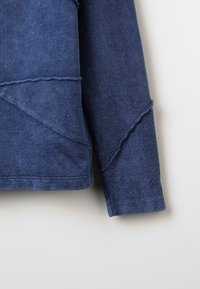 Maglione in denim blu scuro con finitura testurizzata, caratterizzato da un design asimmetrico e bordi grezzi lungo le cuciture e le maniche.