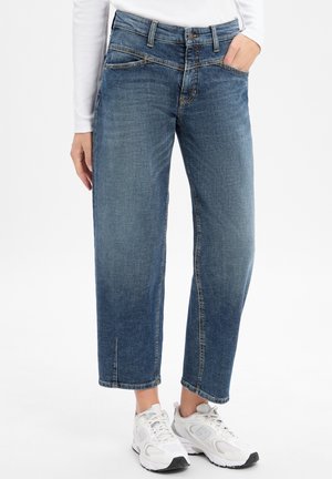 Person wearing cropped blue jeans and white sneakers with one hand in pocket against white background.