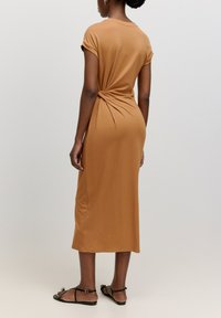 Robe ajustée, longueur genou, en tissu marron clair. Présente un détail de nœud tordu à l'arrière et des manches courtes, assortie de sandales noires.