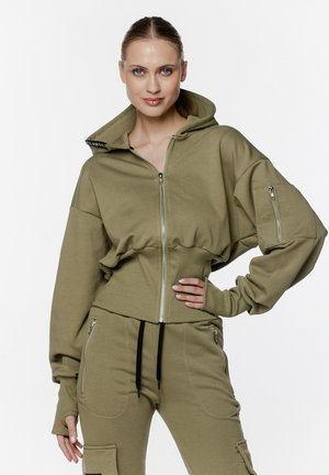 Oliwkowy, krótki hoodie z pełnym zamkiem, długimi rękawami i bocznymi kieszeniami na zamek. Miękki materiał o luźnym kroju i z kapturem.