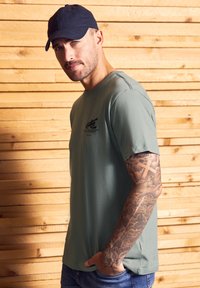 Hombre con el brazo tatuado que lleva una camiseta gris y una gorra negra, de pie de lado contra una pared de tablones de madera con la mano en el bolsillo.
