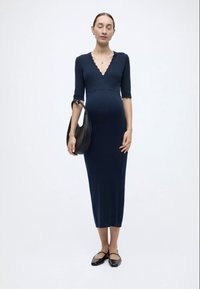 Μπλε σκούρο ριμπ πλεκτό φόρεμα για εγκυμοσύνη με V-λαιμόκοψη, μανίκια τρία τέταρτα και μακριά φούστα. Συνδυασμένο με μαύρη τσάντα ώμου και φλατ παπούτσια.