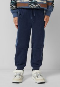 Marineblaue Jogginghosen mit elastischem Bund, Seitentaschen und kontrastierenden blauen Einsätzen. Kombiniert mit weißen Sneakers mit Netzelementen.