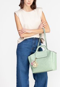 Kvinde i en ærmeløs beige top og blå jeans holder en lys grøn håndtaske med blomsterudskæring og en blomster charms.