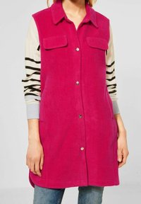Fuchsia ärmlös väst i mjukt tyg, med krage, två framfickor och silvriga knappar, ovanpå en randig långärmad skjorta.