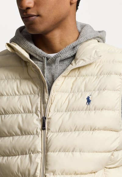 Doudoune crème avec des sections cousues et une fermeture éclair à l'avant. Présente un petit logo bleu au-dessus de la poitrine gauche, superposé à un sweat à capuche gris.