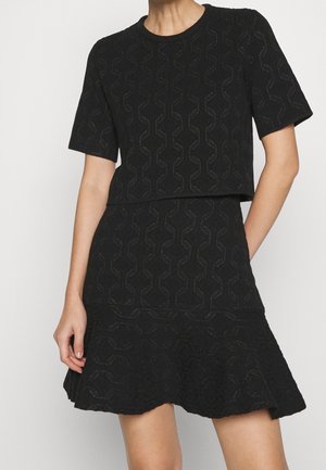 Frau trägt ein schwarzes, strukturiertes Kurzarm-Oberteil und einen passenden ausgestellten Rock mit geometrischen Mustern, steht mit einer entspannten Hand.