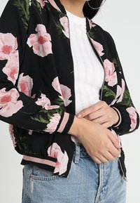 Giacca bomber nera con grandi motivi floreali rosa, polsini e vita a costine; presenta una chiusura con zip sul davanti e un tessuto leggero.