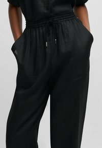 Czarne luźne spodnie wykonane z lekkiego materiału, z elastycznym paskiem w talii z troczkiem, bocznymi kieszeniami i gładką fakturą.