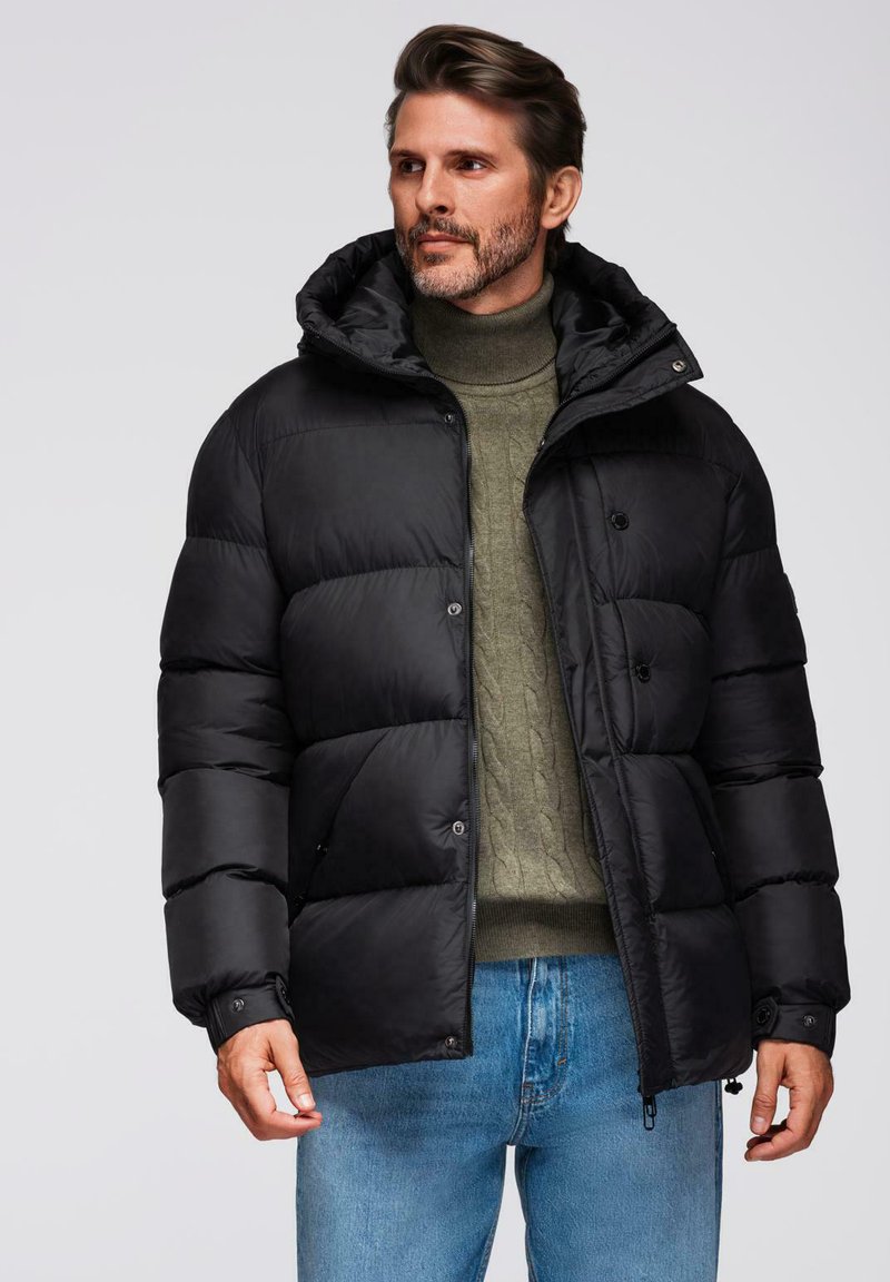Abrigo de plumas negro con capucha, con un patrón acolchado, botones frontales y bolsillos laterales. Combinado con un jersey de lana verde y jeans azules.