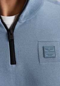 Lichtblauwe zip-up trui met geribde kraag, textuurstof, zwarte rits en een vierkante logo patch op de linkerborst.