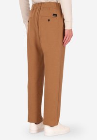 Pantaloni della tuta marroni con una vestibilità rilassata, dotati di un elastico in vita, due tasche posteriori con bottoni e un'etichetta di branding discreta. Tessuto liscio e texture.