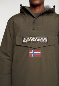 Oliwkowa kurtka z kapturem o designie z półzamek, z dużą przednią kieszenią i naszywkami, w tym logo i norweską flagą.