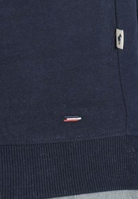 Pull marine en bleu foncé en tissu doux, avec des poignets et un ourlet côtelés. Détail notable : un petit logo tricolore près du bas.
