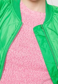 Close-up van een roze gebreide sweater onder een opengeritste groene gewatteerde jas op een persoon.