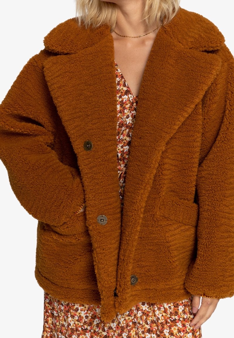 Veste marron, pelucheuse et surdimensionnée avec un grand col, deux poches avant et des boutons noirs. Portée par-dessus une robe à motif floral.