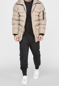 Giacca imbottita beige con cappuccio, chiusura a zip e tasche applicate. Abbinata a pantaloni cargo neri e sneakers bianche per un look casual.