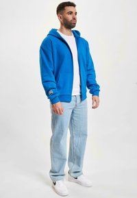 Haut à zip bleu avec une poche kangourou et un détail de logo sur la manche, associé à un jean bleu clair et des baskets blanches.