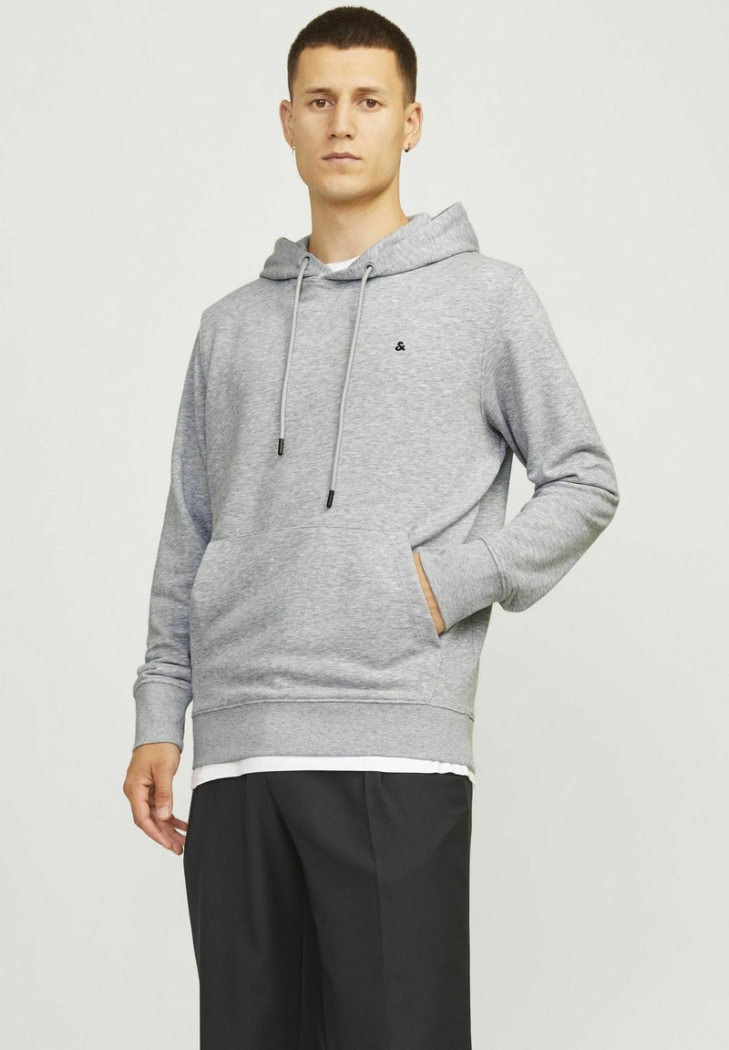 Grauer Hoodie mit Kordeln, Fronttasche und gerippten Bündchen. Mit kleinem schwarzem Logo und geschichtetem Saum über schwarzen Hosen.