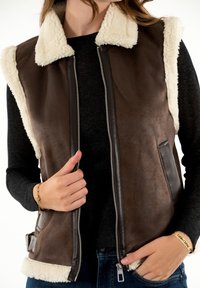 Chaleco de ante sintético marrón con cuello y ribete de sherpa color crema. Cuenta con cierre de cremallera, bolsillos laterales y detalles de cuero. Parece acogedor y cálido.