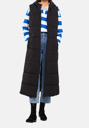 Femme portant une longue veste matelassée noire sans manches par-dessus une chemise à rayures bleues et blanches, un jean bleu, et des bottes noires à lacets.