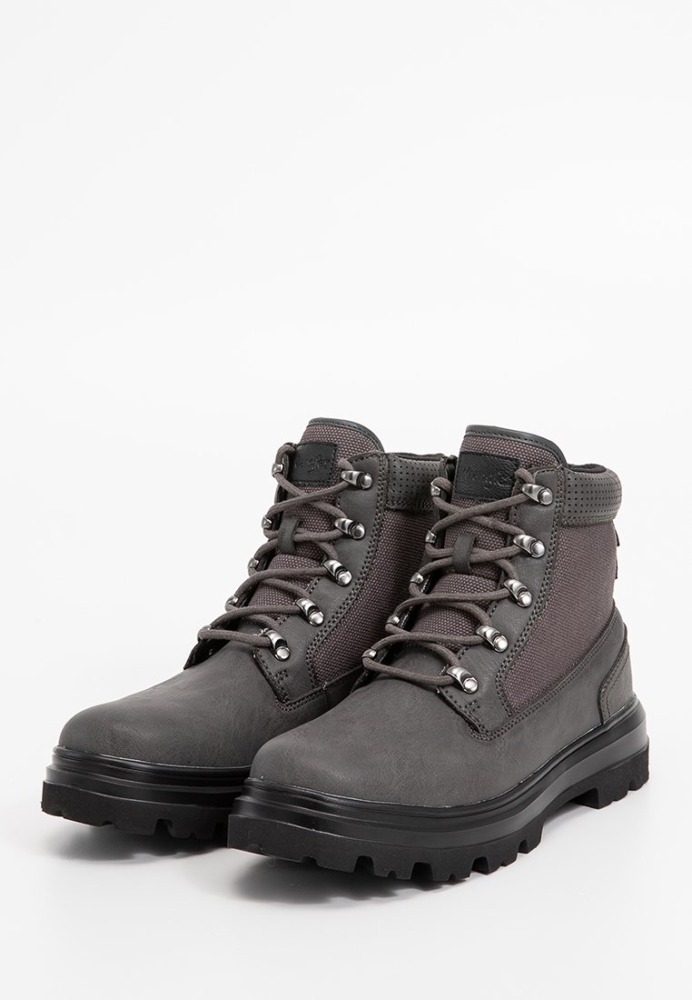 Graue Knöchelstiefel mit einer strukturierten Gummisohle, Metallösen und Schnürsenkeln. Das Obermaterial weist eine Mischung aus glatten und stofflichen Bereichen auf.
