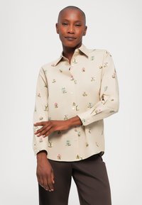 Beige overhemd met knoopsluiting, voorzien van kleurrijke bloemenborduurwerk, lange mouwen, een klassieke kraag en een ontspannen pasvorm. In bruin broek gestoken.