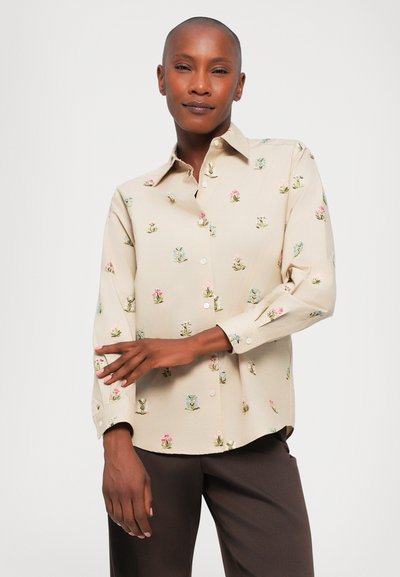 Beżowa koszula zapinana na guziki z kolorowym haftem kwiatowym, długimi rękawami, klasycznym kołnierzykiem i luźnym krojem. Włożona w brązowe spodnie.
